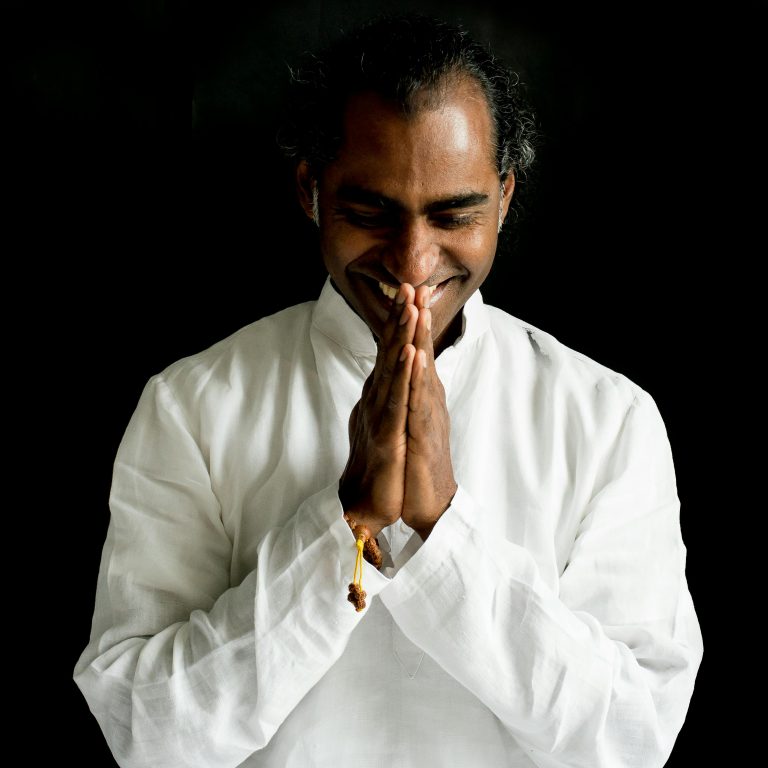 Portrait of a smiling man in white attire with hands in Namaste gesture.